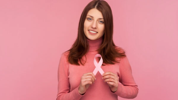 Cute smiling brunette woman in pink sweater holding pink ribbon near her chest supporting another women, international symbol of breast cancer awareness