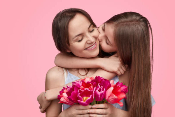 Little girl kissing cheerful woman in cheek presenting bouquet of tulips for spring holiday on pink background