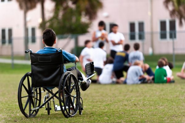 twelve years old child sitting in a wheel chair watching other kids getting together in the park while he is left behind