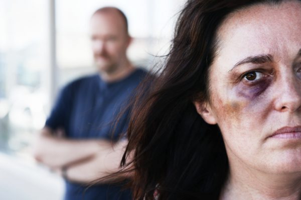 A beautiful but desperate looking woman, bruised and with a black, eye gazes out at camera. Out of focus in the background, a sinister-looking man lurks, probably the perpetrator of the violence.